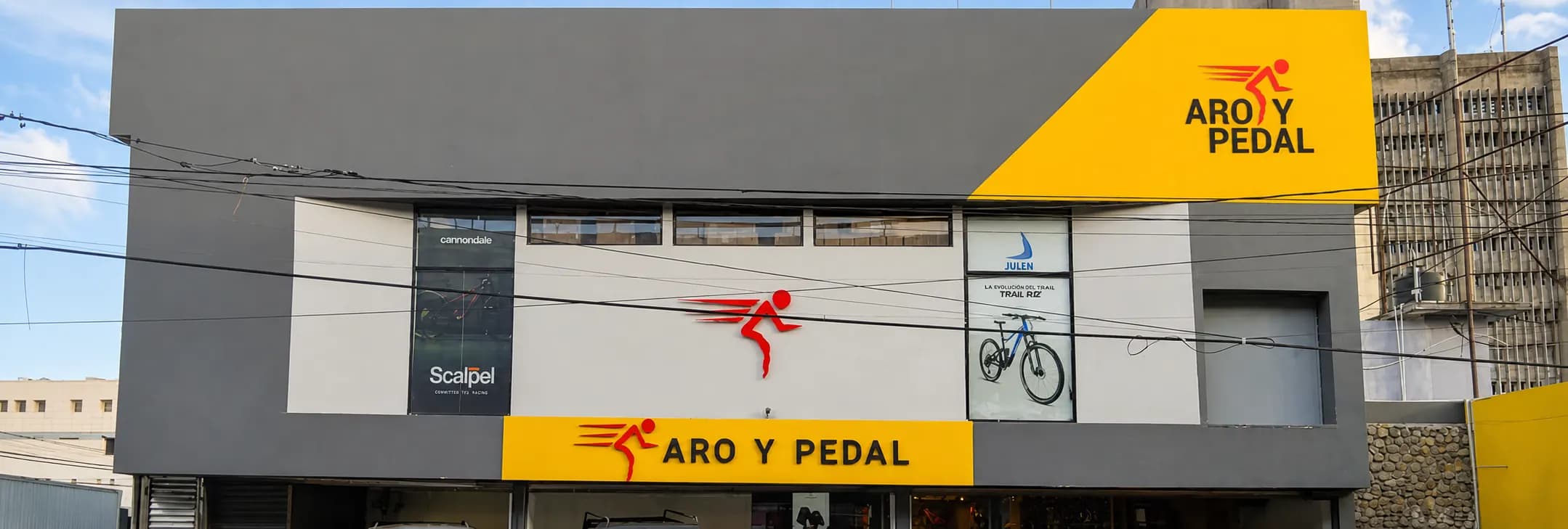 Fachada de la tienda de Aro y Pedal en la avenida 27 de Febrero, Santo Domingo: letrero amarillo con el logo en la esquina y la banda amarilla Aro y Pedal sobre la entrada